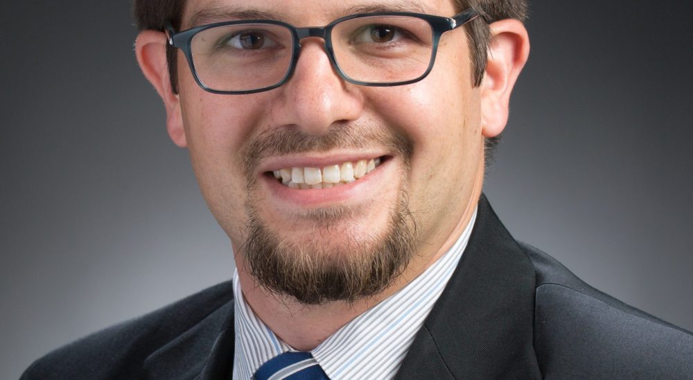 A white millennial man with glasses in a suit and tie posing for a classic portrait with a smile on his face.